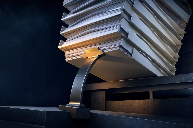 A heavy stack of white process trays and review folders is being held up by a single brass budget tab that has transformed into a bowed steel brace, showing invisible load-bearing work holding a system together.