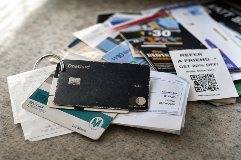 A worn bank card clipped to a transit pass, grocery receipt, and utility bill sits in sharp focus, while a pile of colorful promotional payment cards blurs into the background.